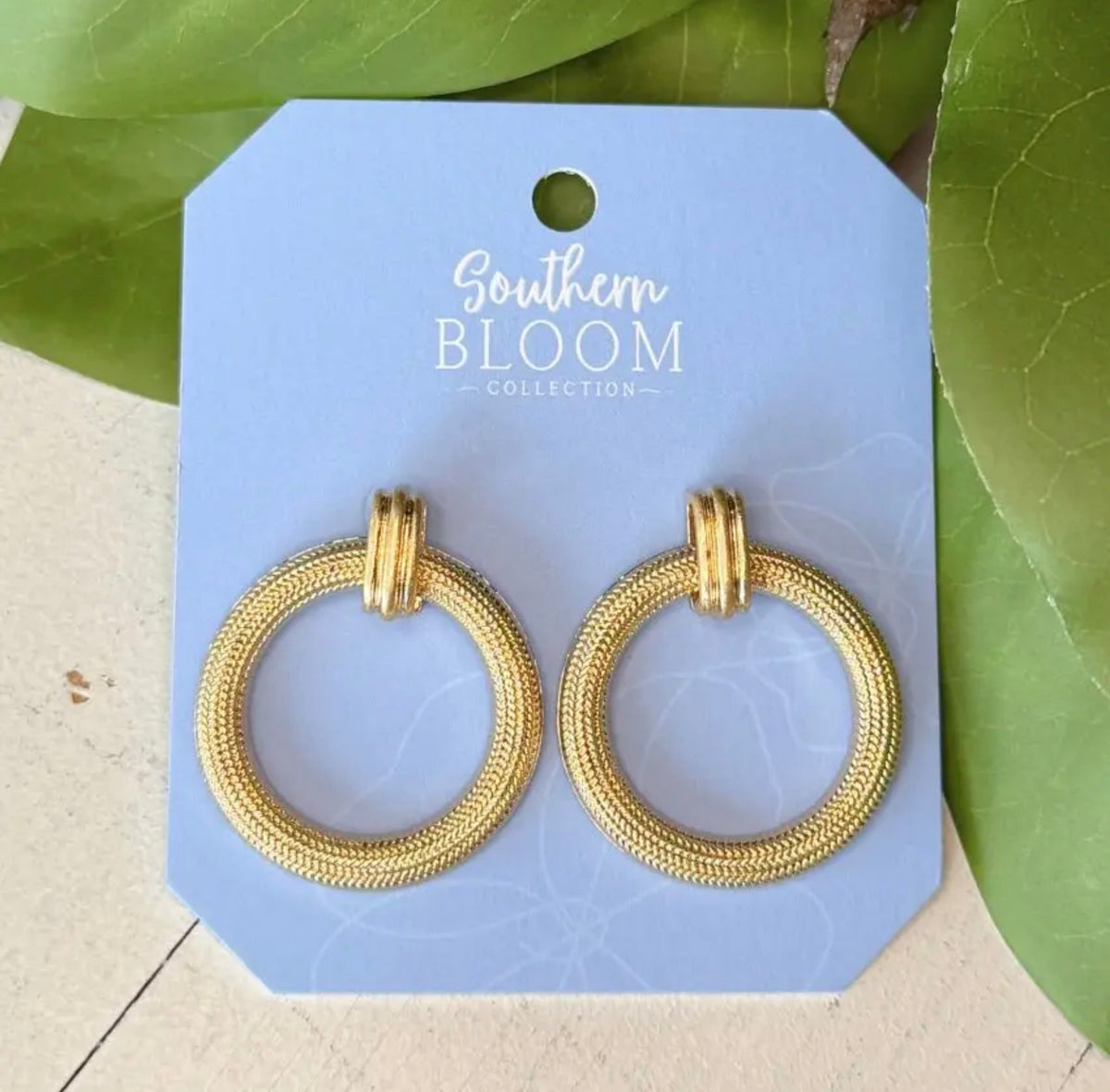 A pair of gold textured hoop earrings on a light blue display card labeled 'Southern Bloom Collection,' photographed with green leaves around it.