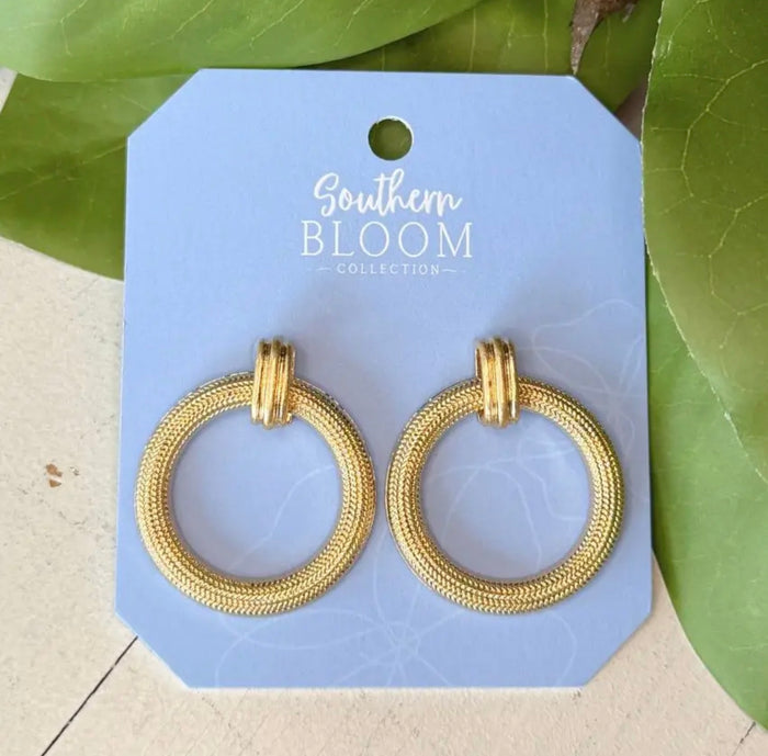 A pair of gold textured hoop earrings on a light blue display card labeled 'Southern Bloom Collection,' photographed with green leaves around it.