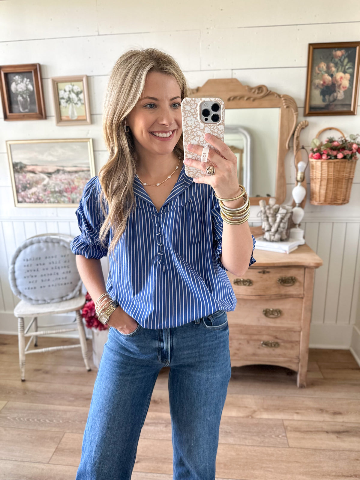 Woman taking a mirror selfie wearing a blue striped shirt and jeans in a room with wooden furniture and decor.