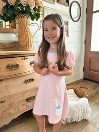 Young girl in a pink dress standing in a room with wooden furniture and decorative items.