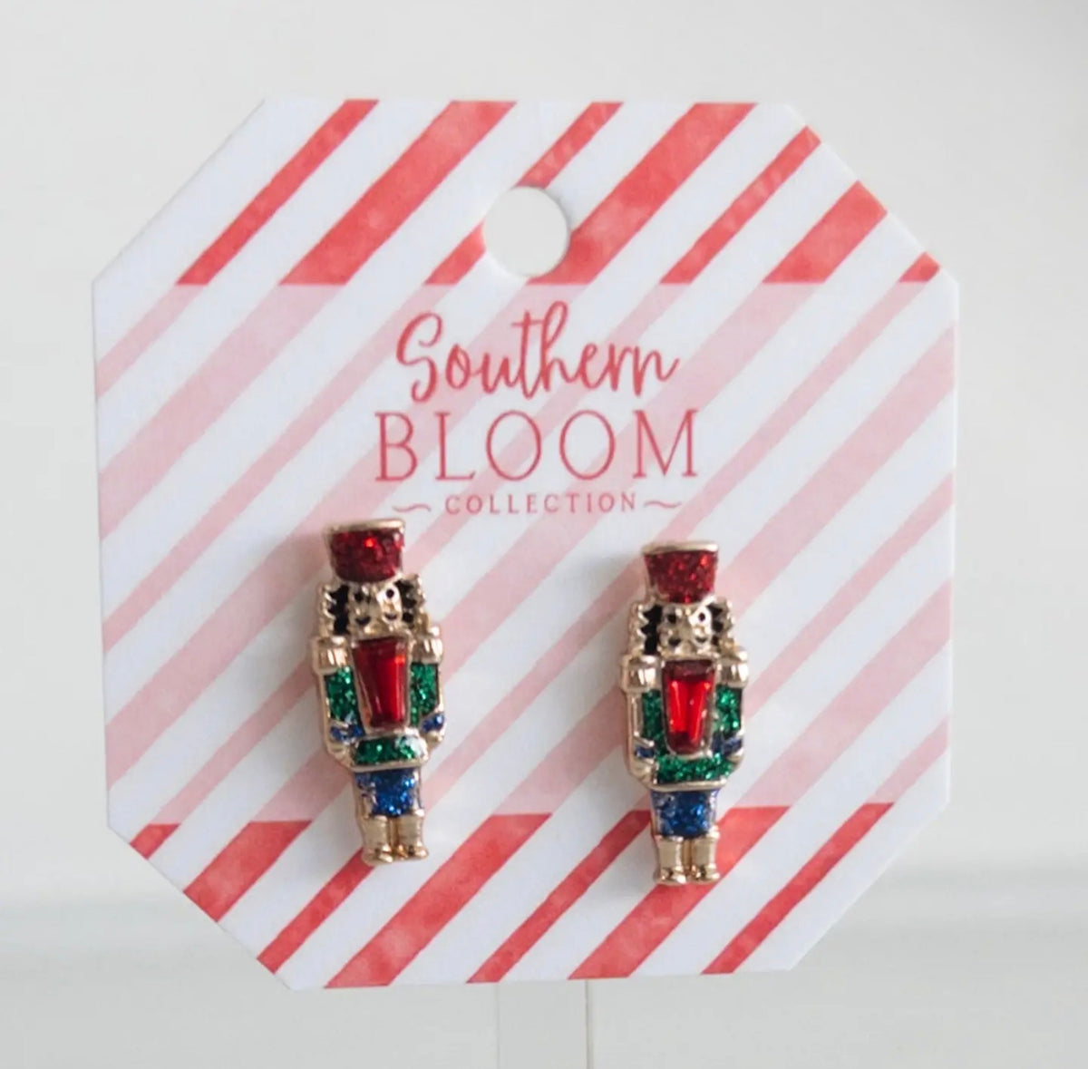 A pair of nutcracker stud earrings from the Southern Bloom Collection, displayed on a red and white striped octagonal backing card. The earrings feature gold-plated metal with red, green, and blue enamel details.