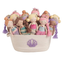 A collection of soft fabric mermaid dolls with colorful dresses and braids, arranged inside a cream-colored woven basket with a purple shell applique, on a solid white background.