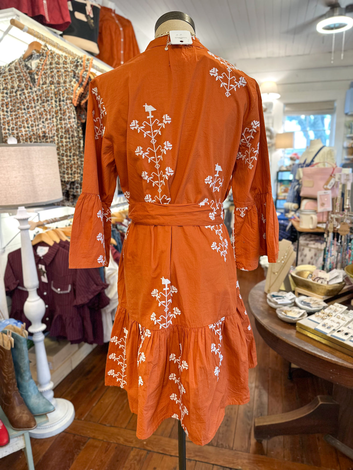 An orange long-sleeve wrap-style dress with white floral embroidery, displayed on a mannequin inside a clothing boutique.