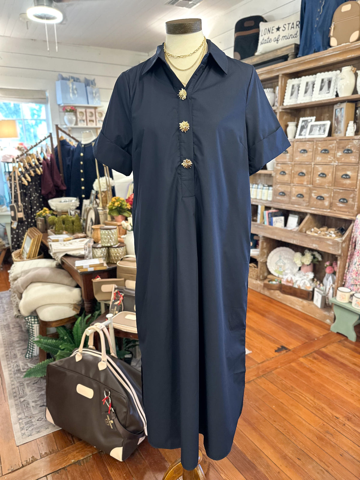 A navy blue short-sleeve collared dress displayed on a mannequin inside a retail shop. The dress has a buttoned chest with decorative gold-toned buttons and is shown against a background of wooden floors, shelves, and clothing racks.