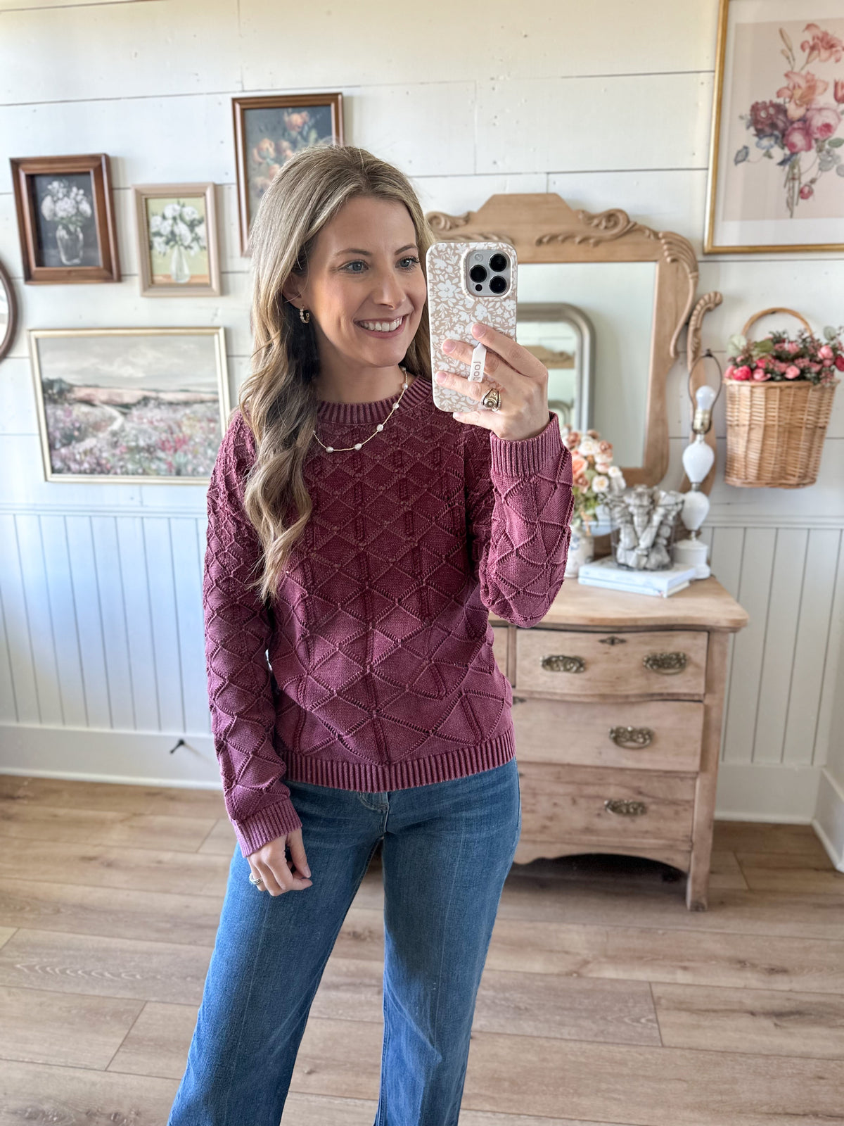 Woman taking a mirror selfie wearing a maroon sweater and blue jeans in a room with wooden furniture and decorative items.