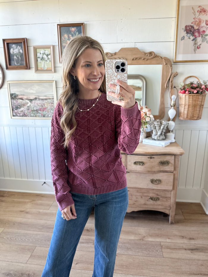 Woman taking a mirror selfie wearing a maroon sweater and blue jeans in a room with wooden furniture and decorative items.