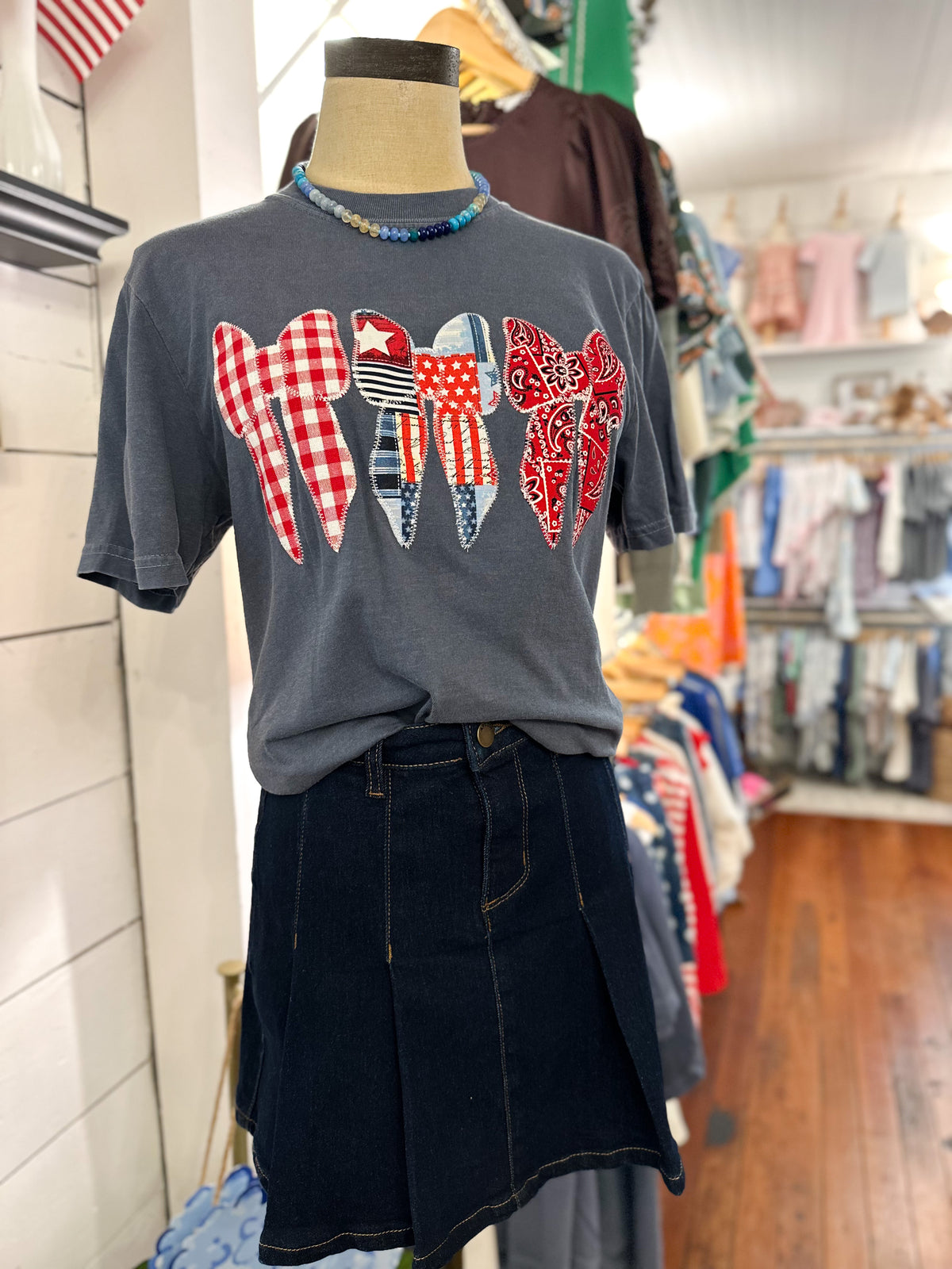 A dark gray graphic t-shirt displayed on a mannequin torso in a clothing boutique. The shirt features a patriotic-themed bow sewn on applique design 