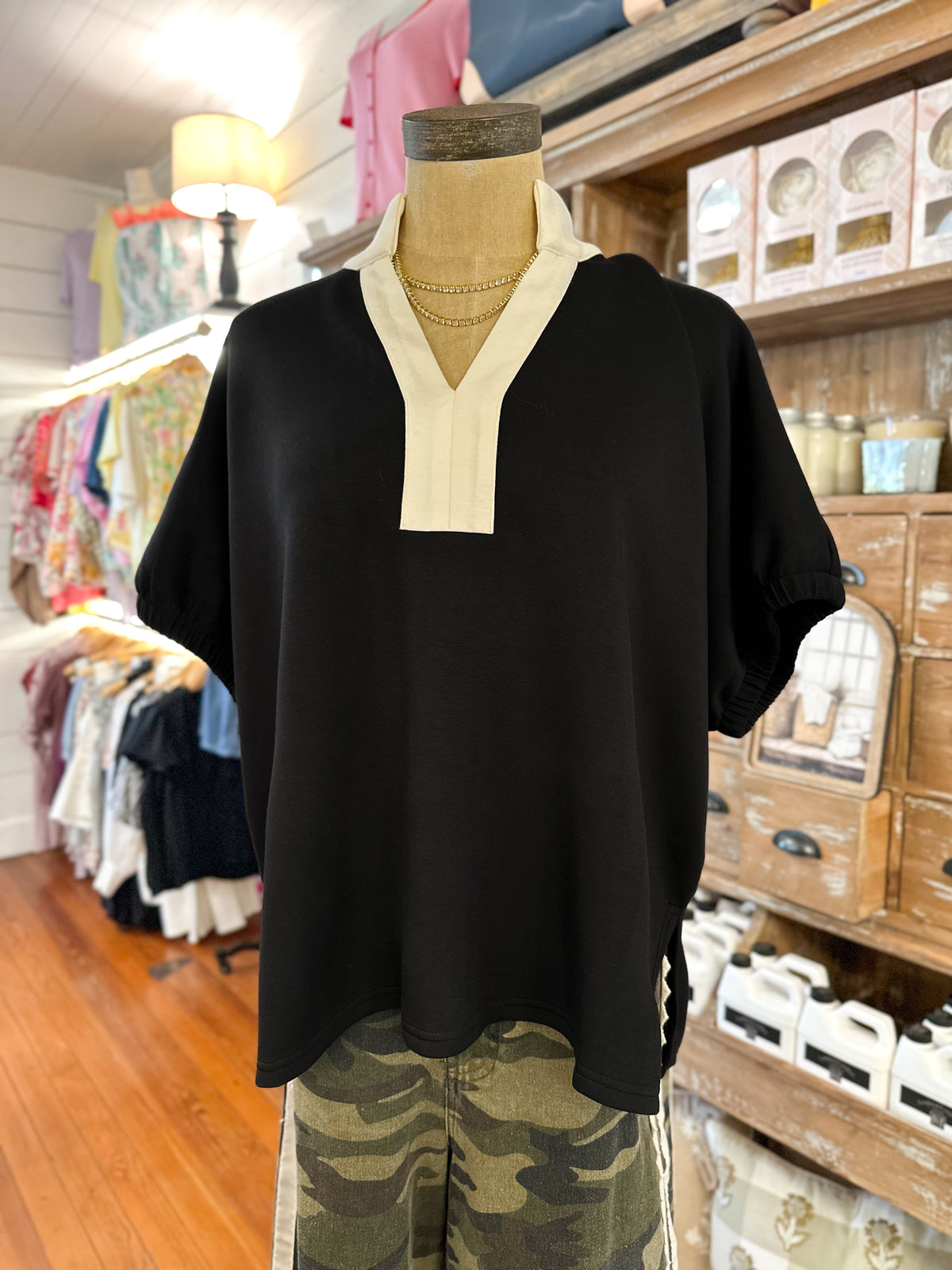 A black short-sleeve top with a cream V-neck collar detail, displayed on a mannequin torso inside a clothing boutique. The mannequin is also styled with camouflage pants.