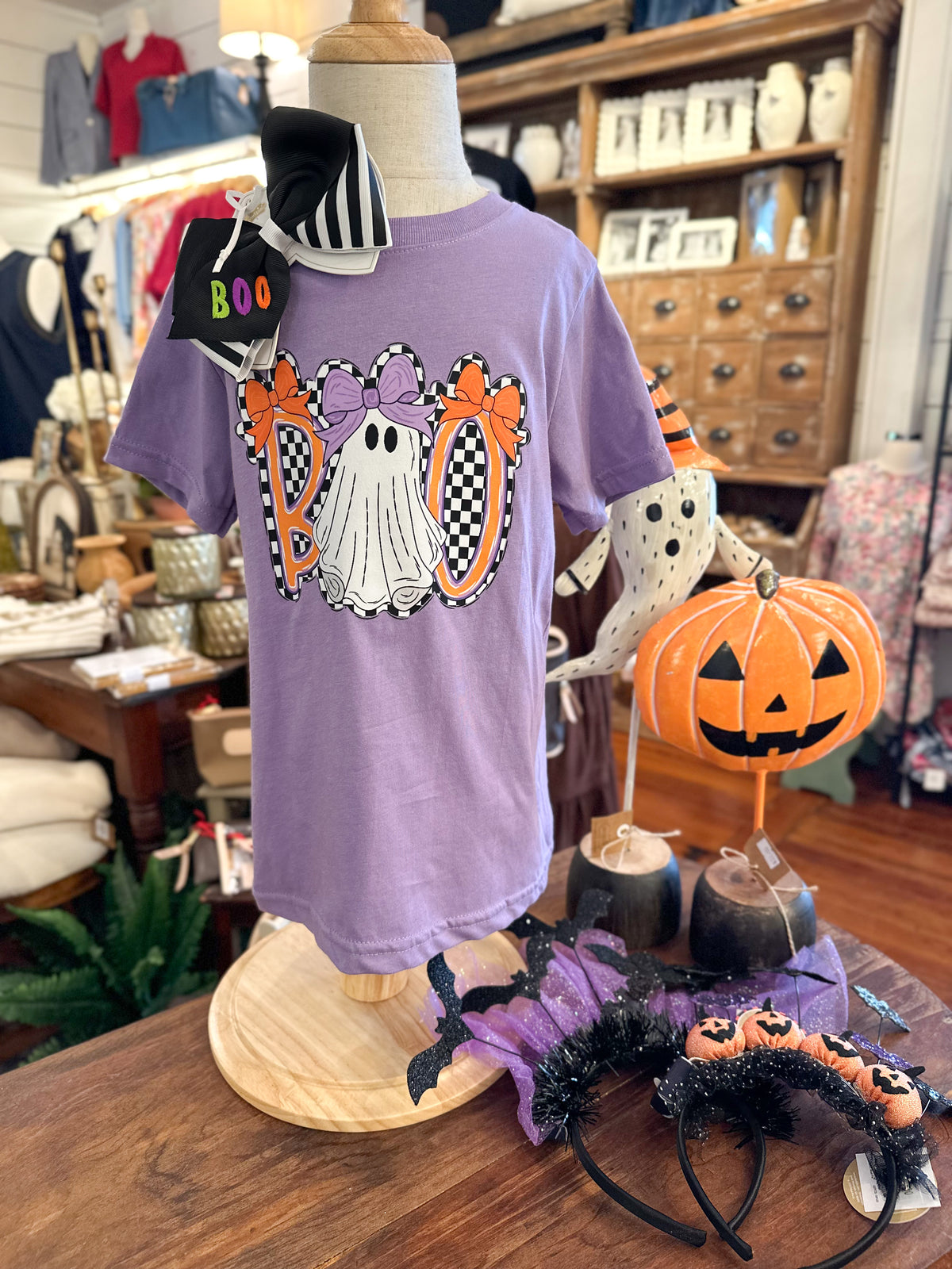 A lavender purple short-sleeve t-shirt displayed on a mannequin torso in a retail shop. The shirt features a Halloween graphic with a white ghost, orange bows, and checkered orange and black shapes.