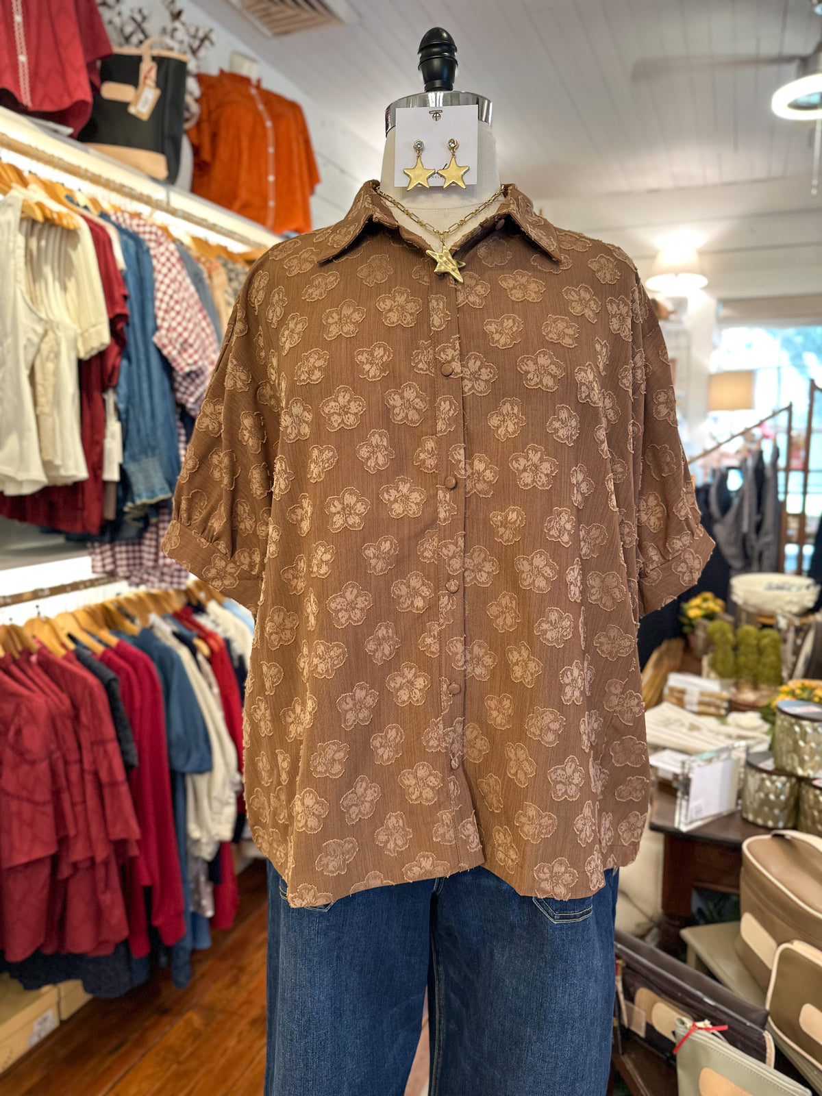 A brown short-sleeve button-up shirt with a floral pattern, displayed on a mannequin torso in a clothing boutique alongside blue jeans.