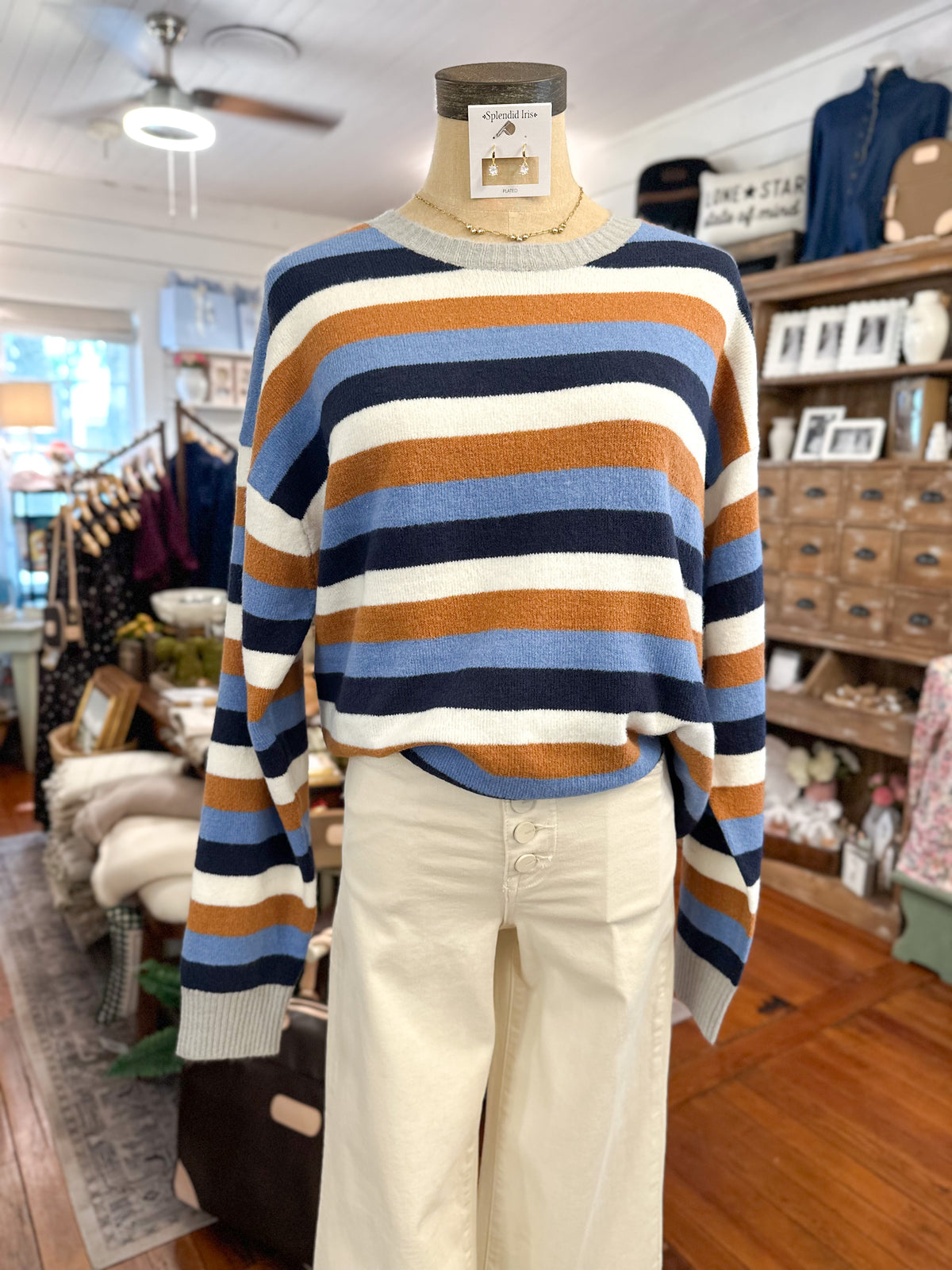A striped knit sweater displayed on a mannequin torso inside a boutique. The sweater has horizontal stripes in cream, navy, light blue, and rust orange.