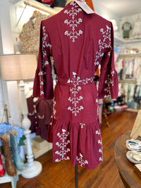 A burgundy long-sleeve mini dress with white floral embroidery, displayed on a mannequin in a boutique setting.