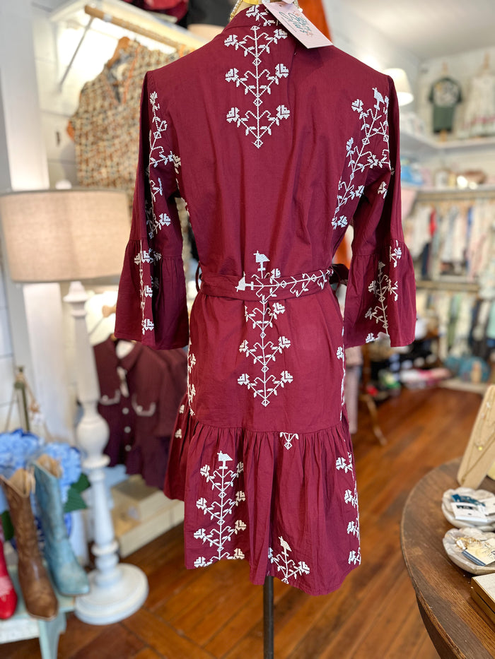 A burgundy long-sleeve mini dress with white floral embroidery, displayed on a mannequin in a boutique setting.