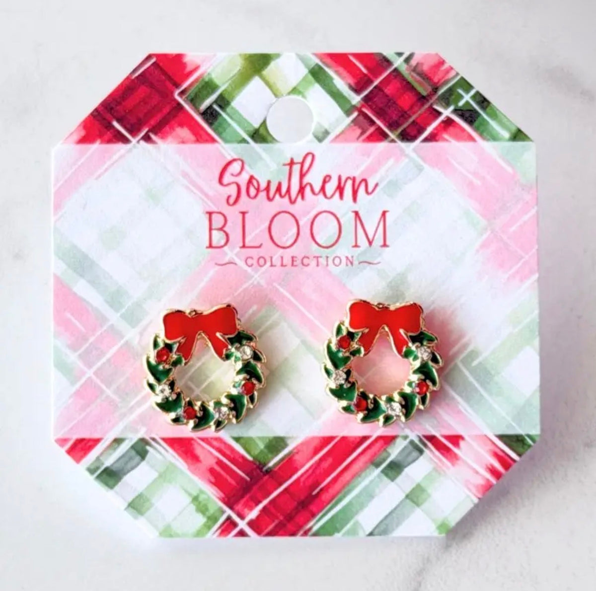 A pair of holiday wreath stud earrings with red bows and green holly details, displayed on a branded card from the Southern Bloom Collection. The card features a festive red, green, and white plaid pattern.