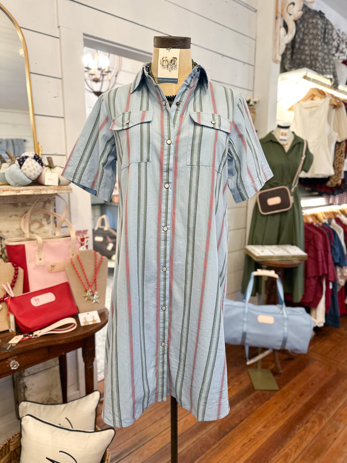 A short-sleeve button-front shirt dress with vertical stripes in light blue, sage green, and pink, displayed on a mannequin inside a boutique.