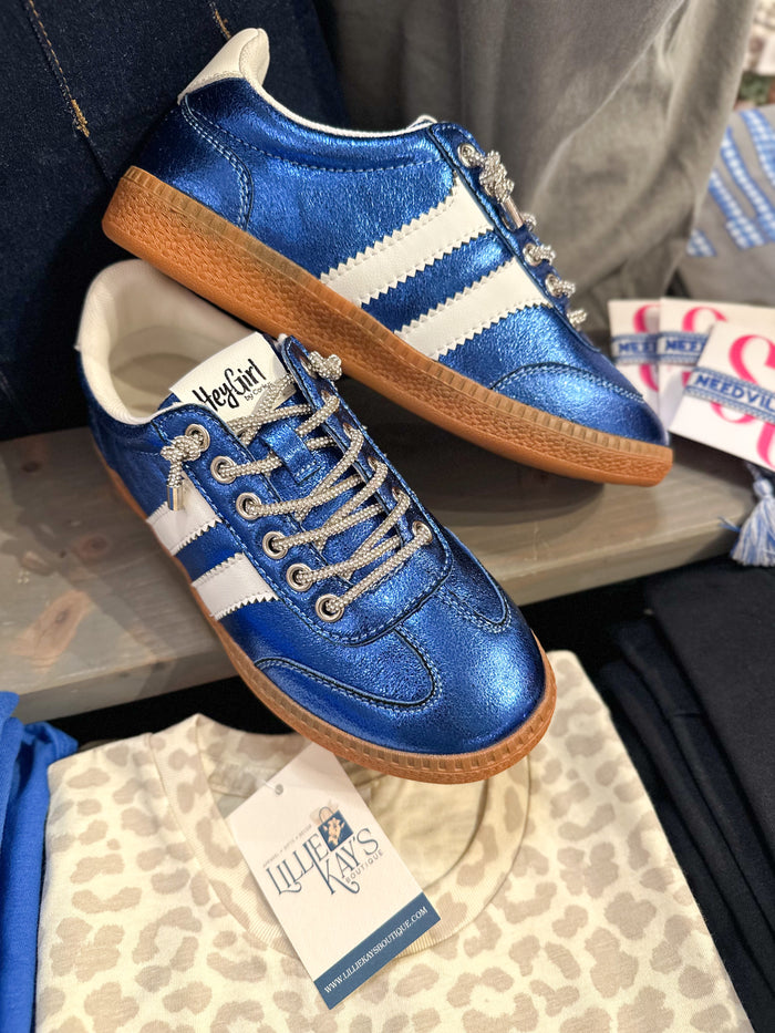 A pair of metallic blue and white low-top sneakers with brown gum soles, displayed on a shelf next to a folded cream leopard-print shirt with a visible 'Lillie' Kay's' tag.