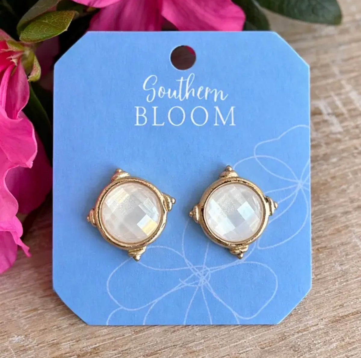 A pair of gold-toned stud earrings with oval milky white  stones, displayed on a blue card labeled Southern Bloom, set against a wooden surface with pink flowers in the background.