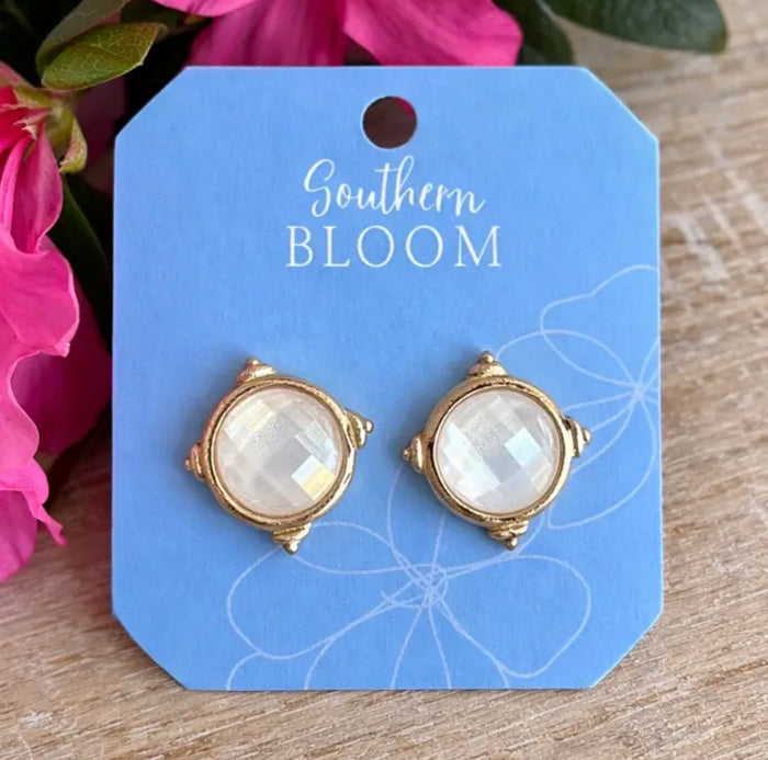 A pair of gold-toned stud earrings with oval milky white  stones, displayed on a blue card labeled Southern Bloom, set against a wooden surface with pink flowers in the background.