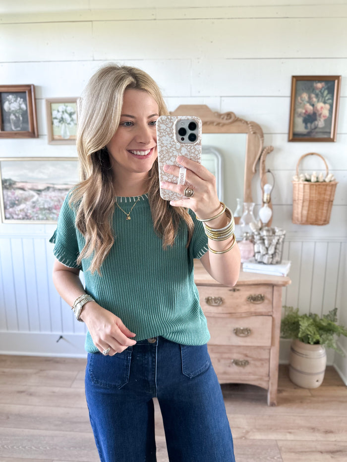 Woman taking a mirror selfie wearing a green sweater and blue jeans in a room with wooden floors and framed pictures on the wall.