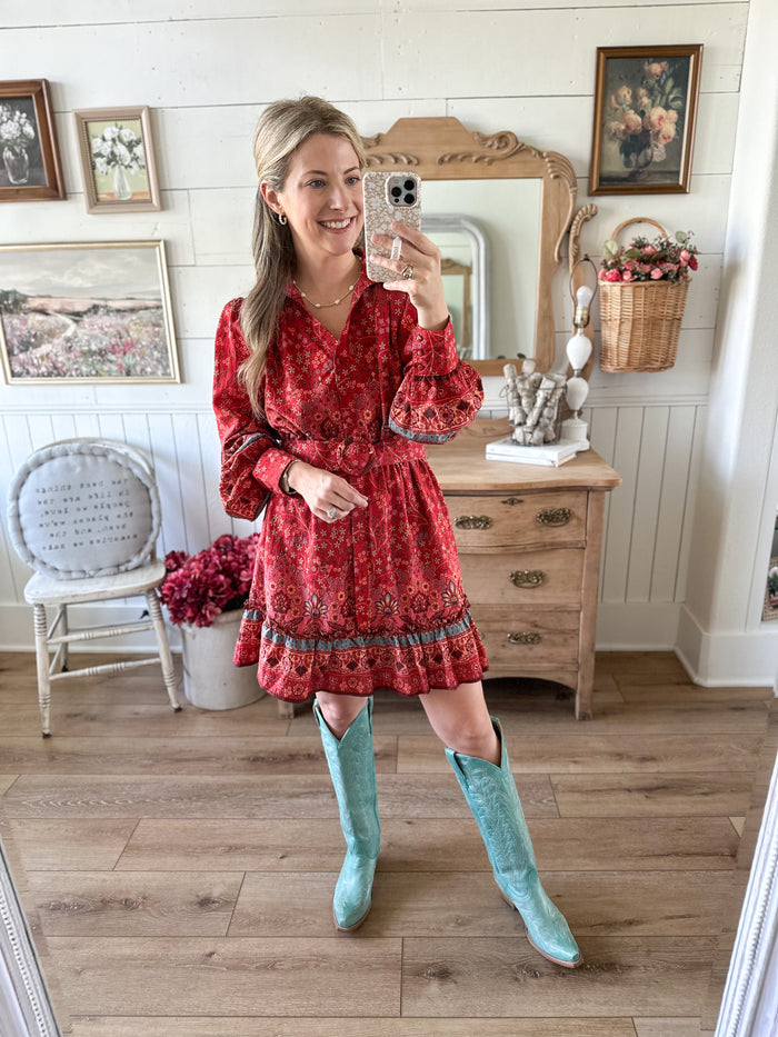 Woman in a red dress with a pattern, wearing turquoise knee-high boots, standing in a room with wooden floors and white walls.