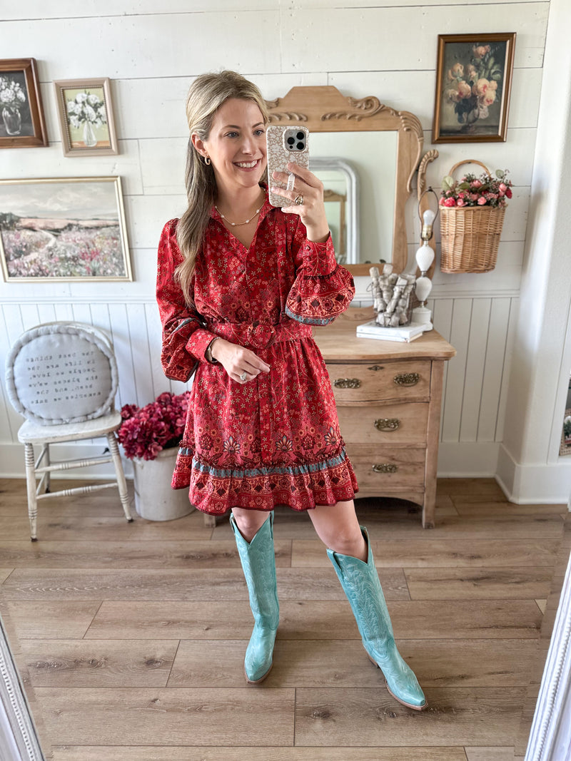 Woman in a red dress with a pattern, wearing turquoise knee-high boots, standing in a room with wooden floors and white walls.