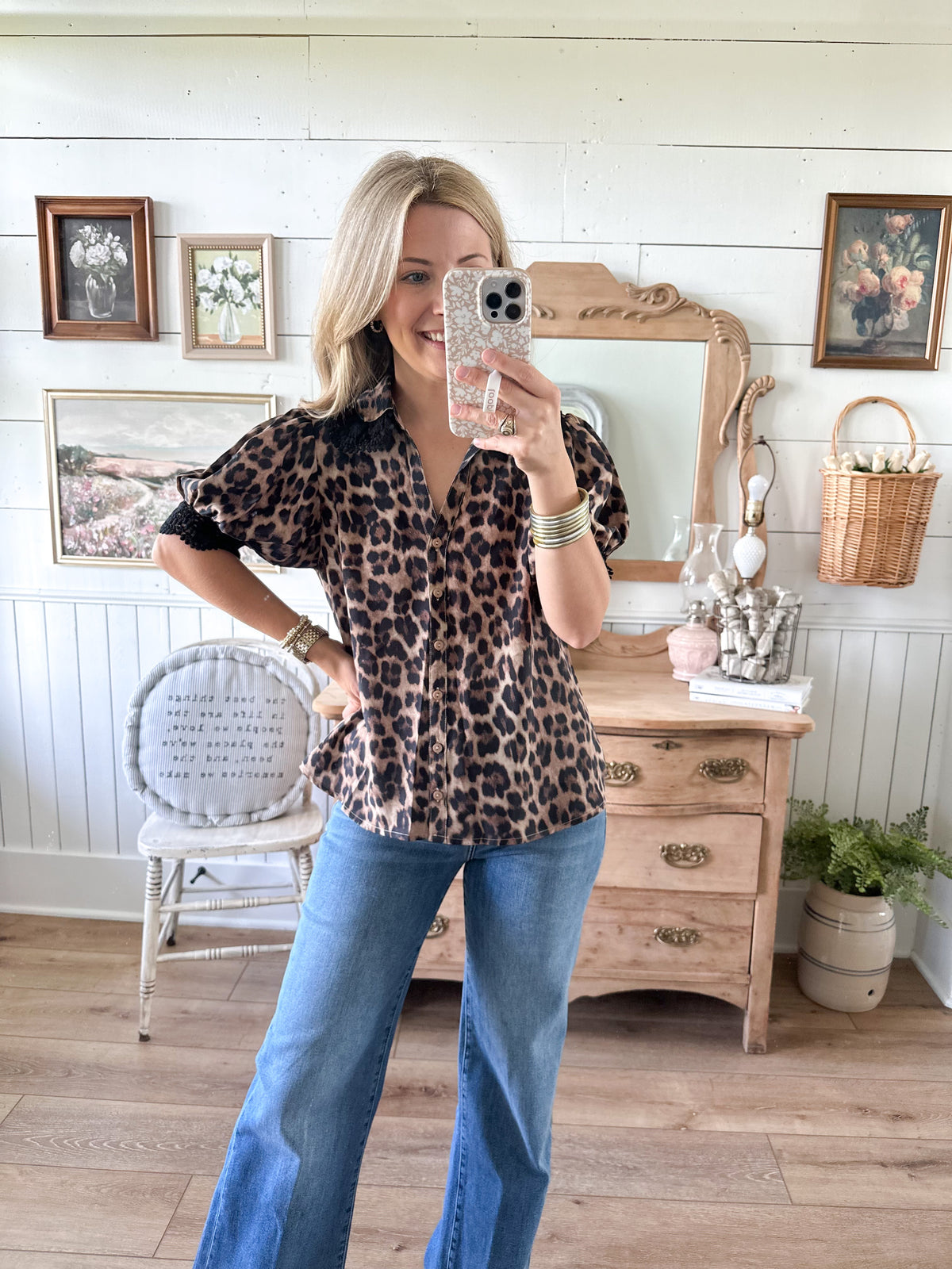 Woman taking a mirror selfie wearing a leopard print shirt and blue jeans in a room with wooden furniture and decor.