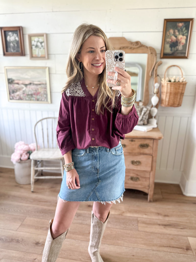 Woman taking a mirror selfie wearing a purple blouse with lace collar, denim skirt, and white boots in a home setting.