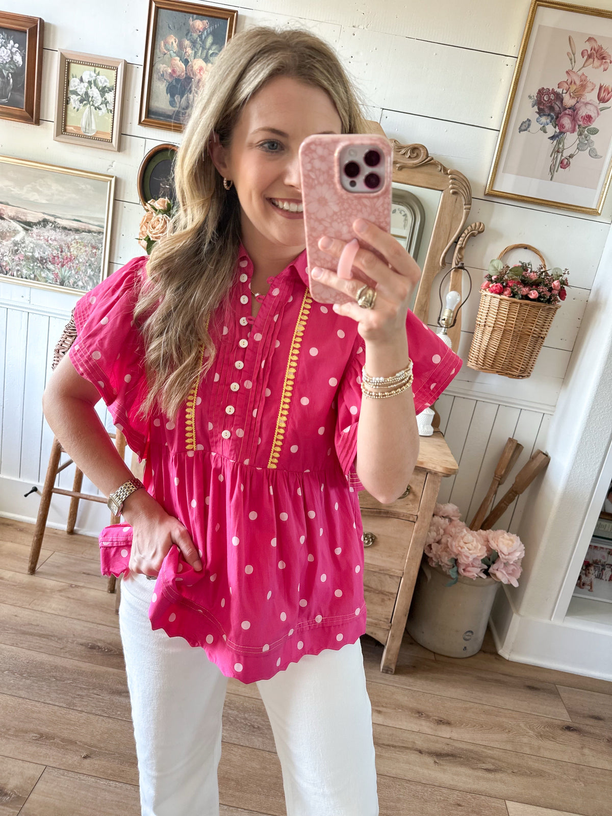 Woman taking a mirror selfie wearing a pink polka dot blouse and white pants in a home setting.