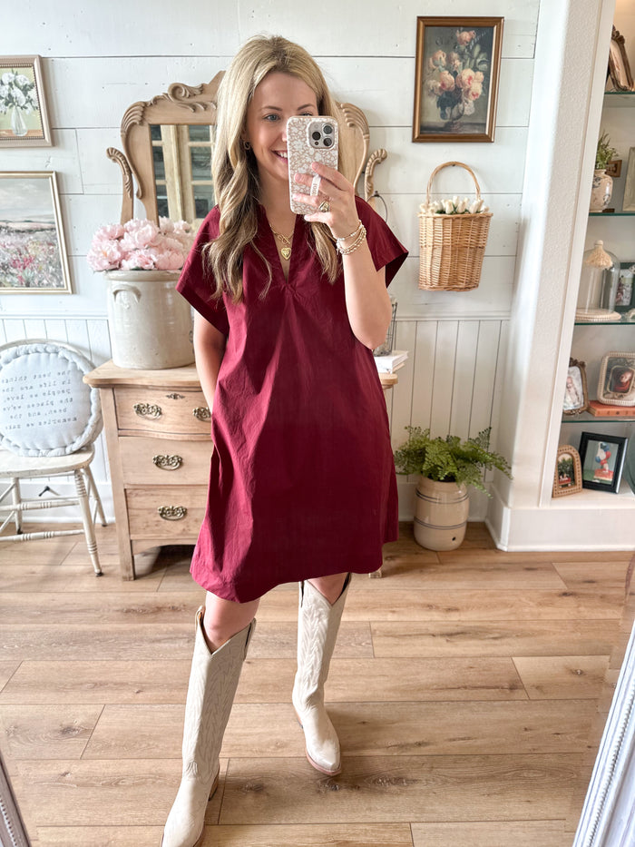 Woman taking a mirror selfie wearing a burgundy dress and white boots in a home setting.