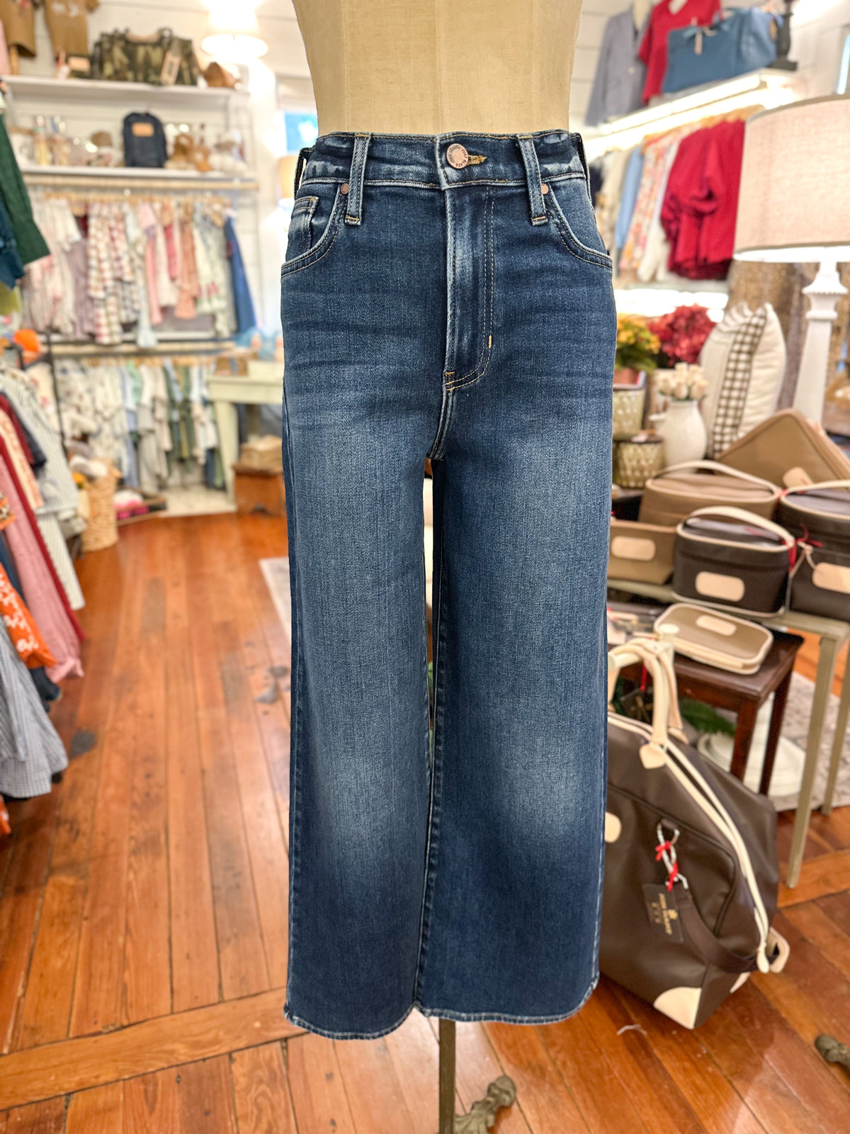 A pair of dark blue wide-leg cropped jeans displayed on a mannequin torso inside a clothing boutique. The background shows wooden floors, racks of other garments, and store fixtures.