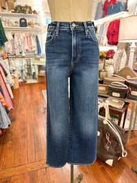 A pair of dark blue wide-leg cropped jeans displayed on a mannequin torso inside a clothing boutique. The background shows wooden floors, racks of other garments, and store fixtures.