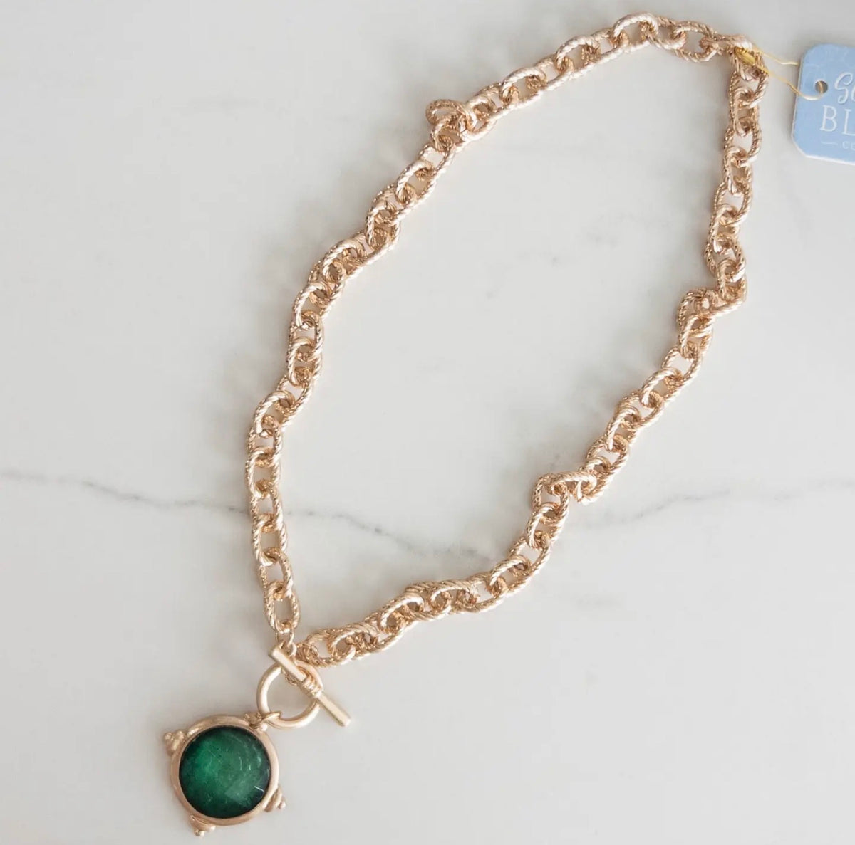 A rose gold chain necklace with a toggle clasp and a round green stone pendant, displayed on a light marble surface.