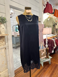 A sleeveless navy satin-like dress with a lace hem, displayed on a mannequin in a boutique setting. The dress features a subtle sheen and a delicate navy lace trim along the bottom edge.