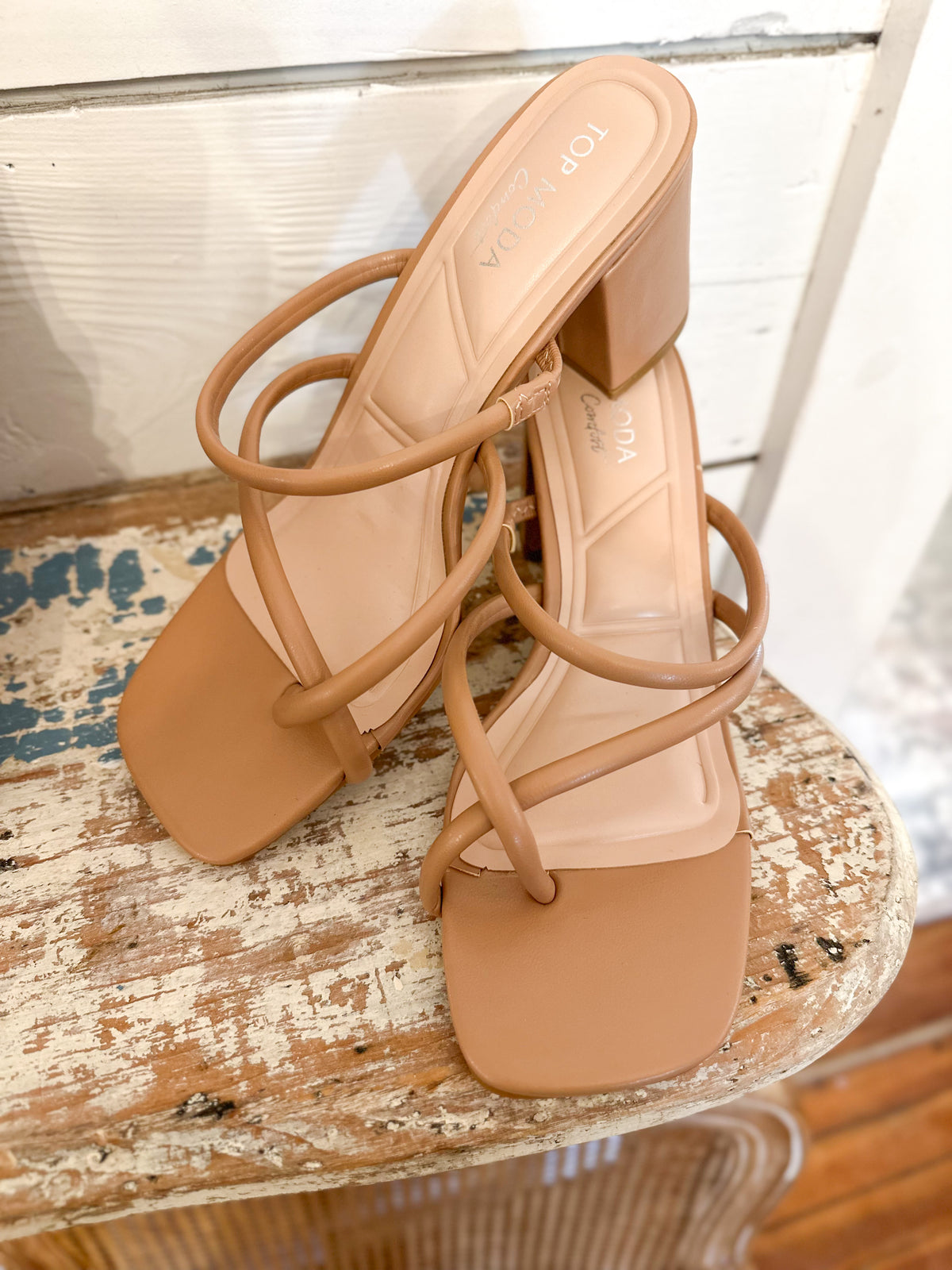 A pair of tan block-heel slide sandals with thin wrap-around straps, displayed on a distressed wooden stool.