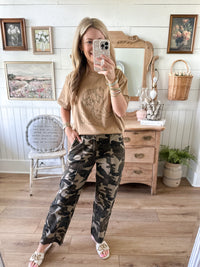 Woman taking a mirror selfie wearing a beige sweater and camouflage pants in a room with wooden furniture and decorative items.