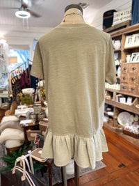 A short-sleeve top in a light Sage green color, displayed on a mannequin from the back in a retail store. The top features a solid upper back panel and a textured, ruffled hem.