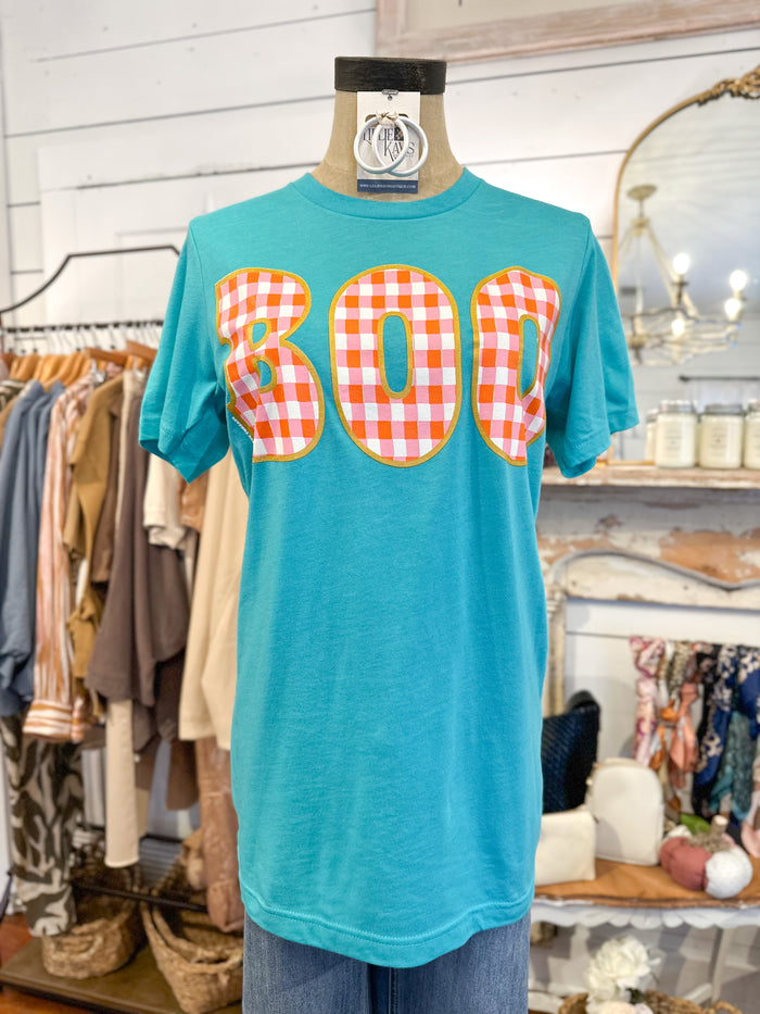 Turquoise t-shirt with 'BOO' in orange checkered pattern on a mannequin in a store setting.
