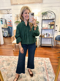 Woman taking a mirror selfie wearing a green top and blue jeans in a room with wooden floors and furniture. green layerz kathleen top