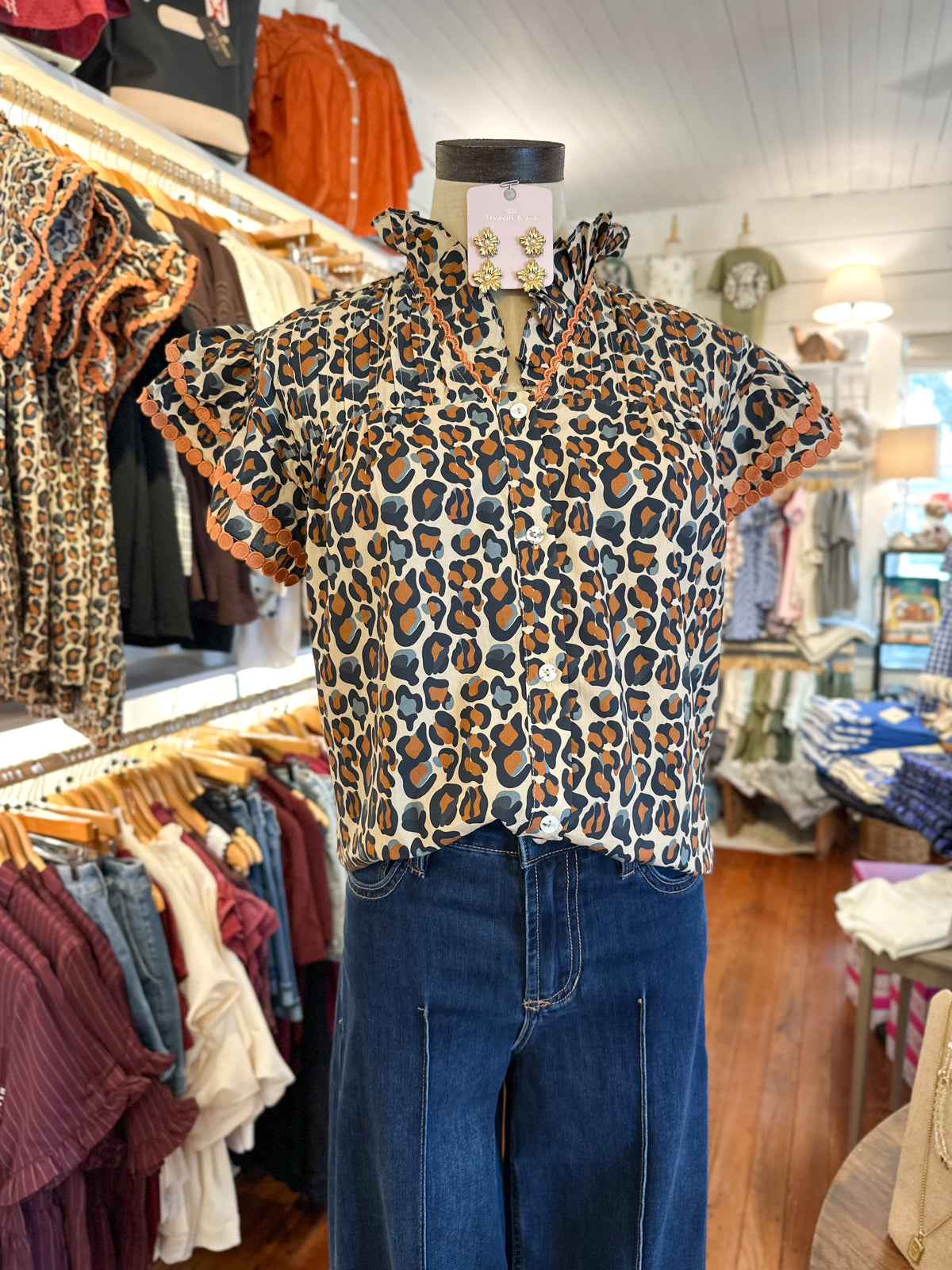 A short-sleeve leopard print blouse displayed on a mannequin torso in a clothing boutique, paired with blue jeans. The blouse features a cream base with brown, black, and orange leopard spots and ruffled trim along the collar and sleeve edges.