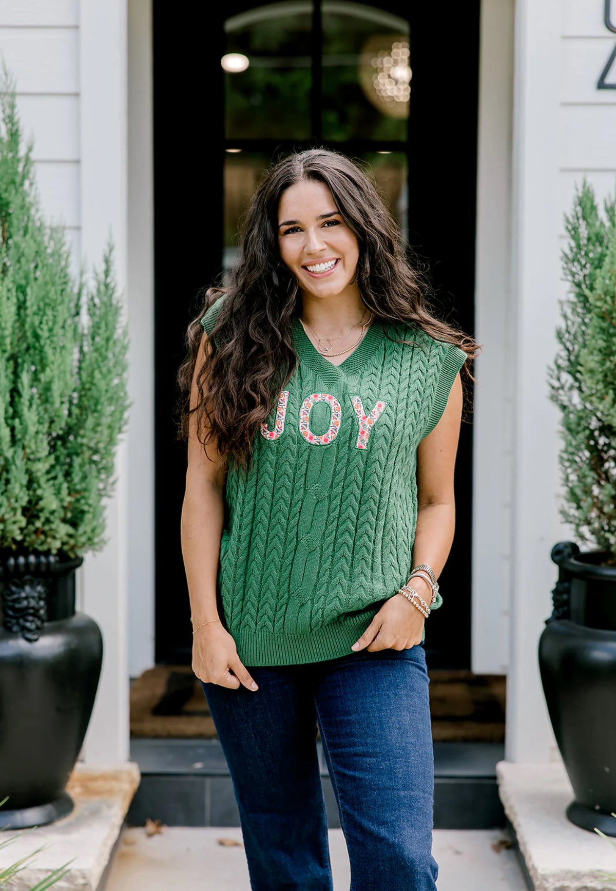 Woman wearing a green sweater with 'JOY' on it, standing in front of a house entrance.