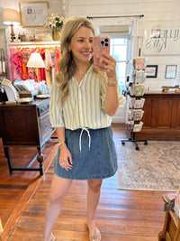 A woman takes a mirror selfie in a boutique shop, wearing a cream and green vertically striped short-sleeve blouse with a drawstring waist and a blue denim skort
