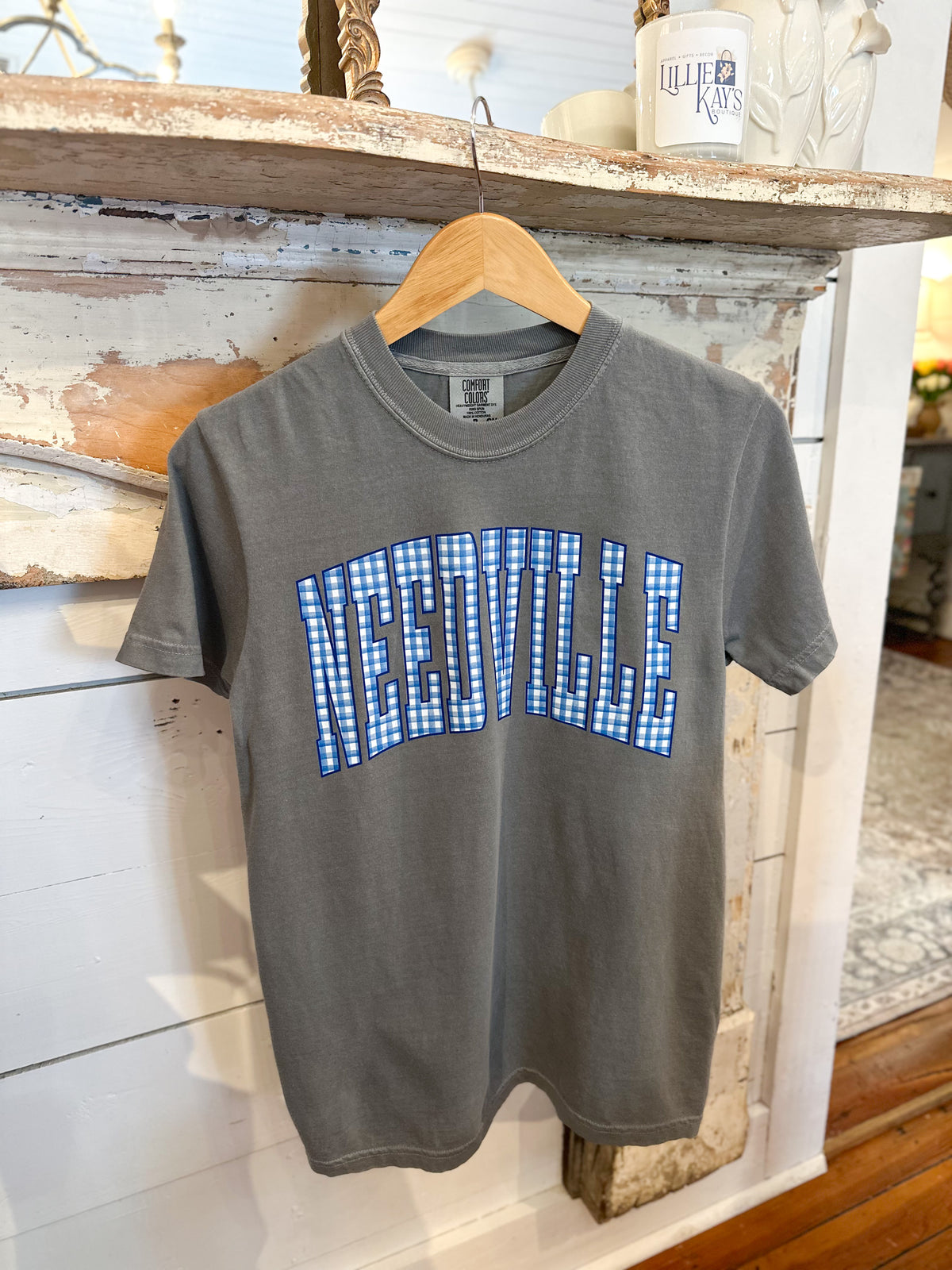 A charcoal gray short-sleeve t-shirt hanging on a wooden hanger against a rustic white wood-paneled wall. The shirt features the word 'NEEDVILLE' printed in an arched, varsity-style font across the chest, filled with a blue and white gingham checkered pattern.