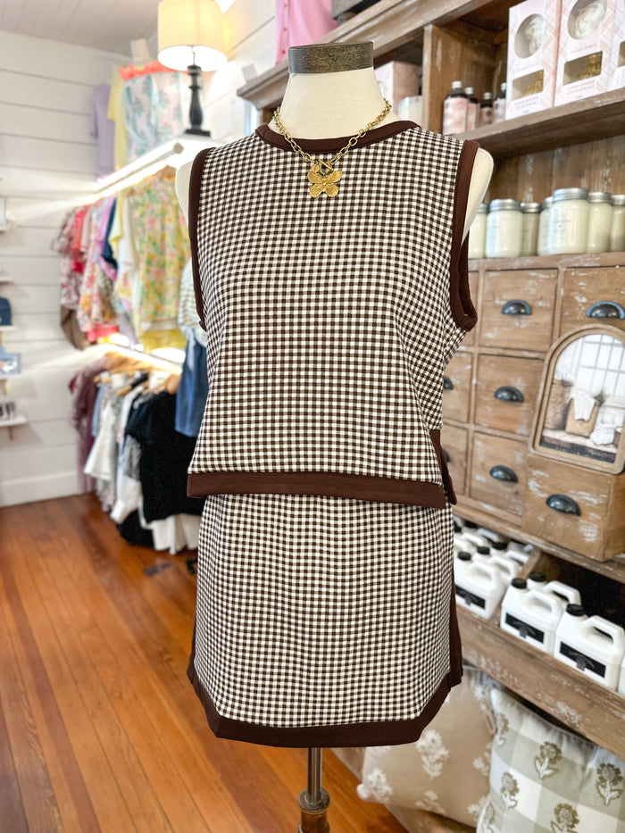 A sleeveless brown and beige gingham dress displayed on a mannequin in a boutique setting, featuring dark brown trim and a gold pendant necklace.