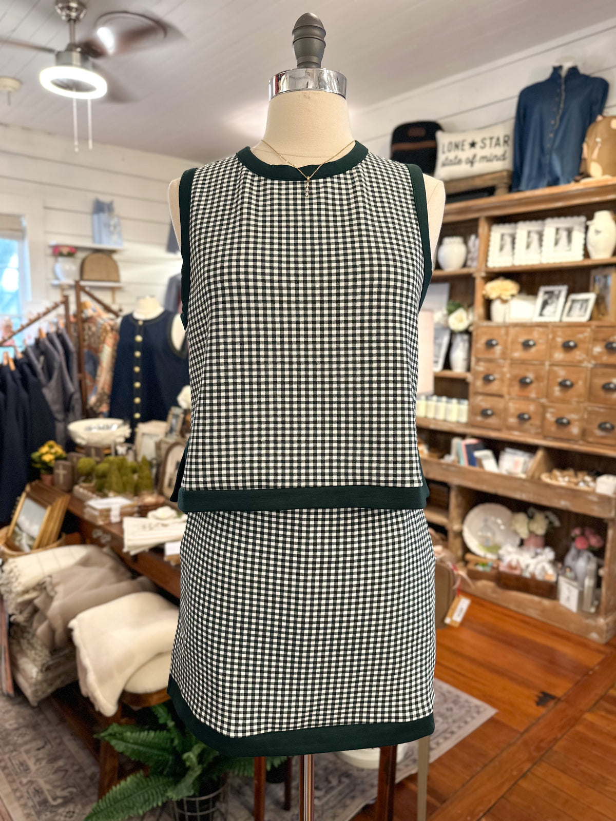 A sleeveless green and white gingham dress displayed on a mannequin inside a boutique. The dress features dark green ribbed trim at the crew neck, armholes, waist, and hem.