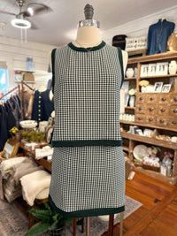 A sleeveless green and white gingham dress displayed on a mannequin inside a boutique. The dress features dark green ribbed trim at the crew neck, armholes, waist, and hem.