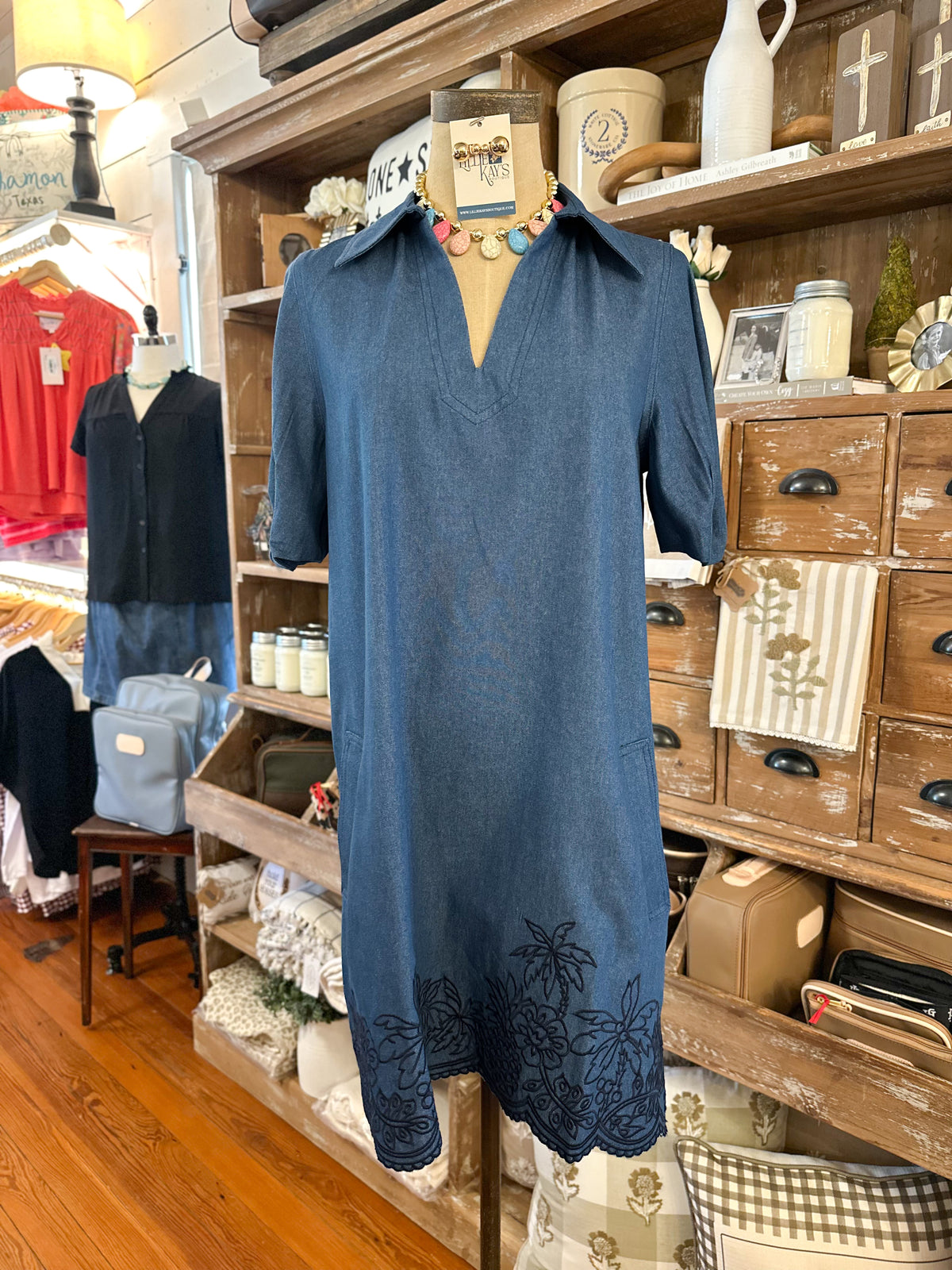 A blue short-sleeve dress displayed on a mannequin torso in a rustic shop interior with wooden shelves and drawers in the background.