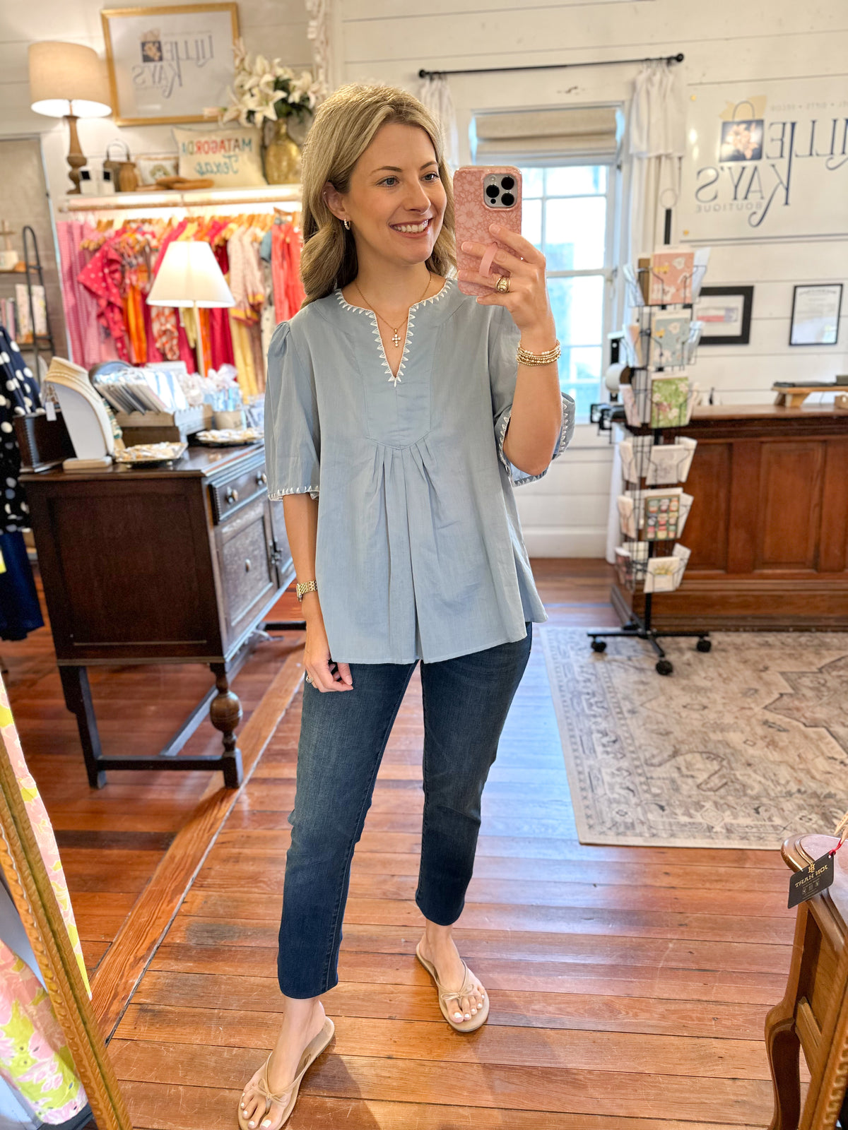 A woman takes a mirror selfie in a boutique while wearing a light blue short-sleeve peplum top with a scalloped neckline, paired with dark blue jeans and beige flat sandals.
