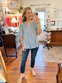 A woman takes a mirror selfie in a boutique while wearing a light blue short-sleeve peplum top with a scalloped neckline, paired with dark blue jeans and beige flat sandals.