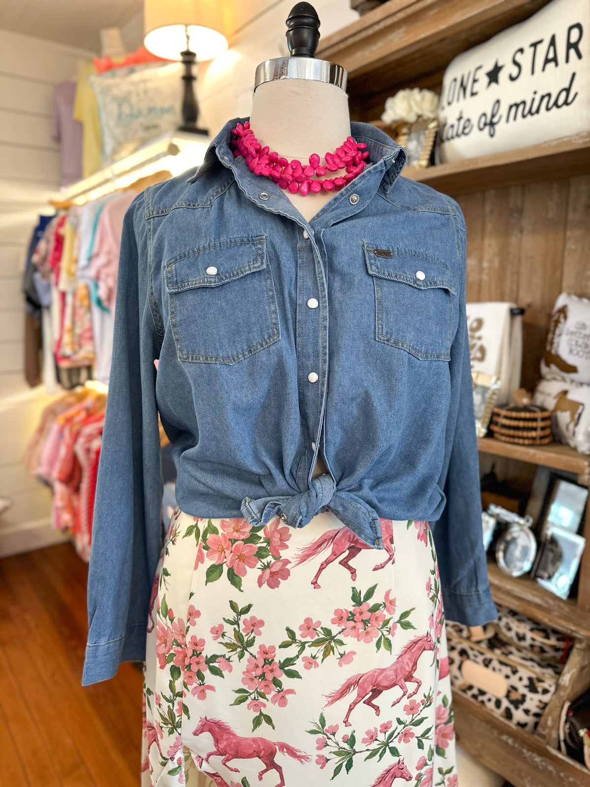 A blue denim button-up shirt with chest flap pockets and white buttons, styled on a mannequin with a bright pink chunky necklace and a cream floral skirt featuring pink horses and flowers. The mannequin is set inside a retail shop with wooden shelves and clothing racks in the background.