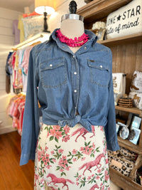 A blue denim button-up shirt with chest flap pockets and white buttons, styled on a mannequin with a bright pink chunky necklace and a cream floral skirt featuring pink horses and flowers. The mannequin is set inside a retail shop with wooden shelves and clothing racks in the background.